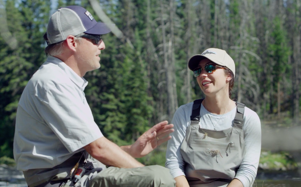 Two people, Derek Bird and April Vokey, standing outdoors with fishing gear and a river in the background.